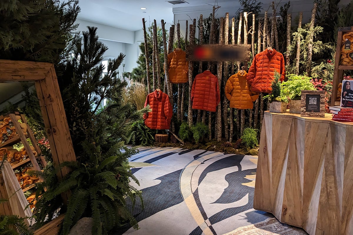 Exposition intérieure sur le thème de la forêt, avec des gilets gonflables orange suspendus à des branches de bouleau, entourés de fougères et de plantes.