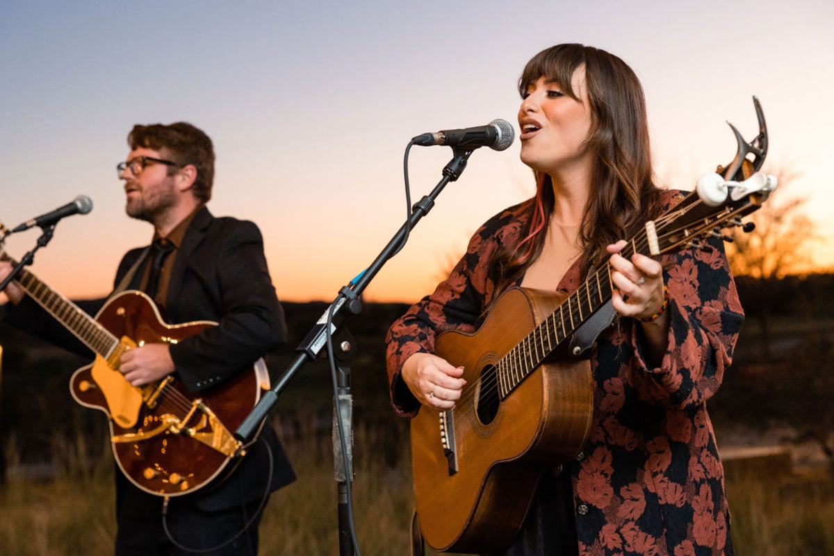 Une chanteuse avec une guitare acoustique et un guitariste se produisent lors d'un concert en plein air au coucher du soleil avec des microphones.