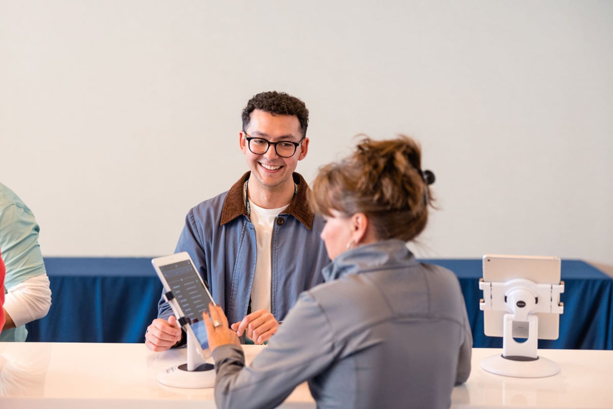 Un employé souriant portant des lunettes aide un client à utiliser sa tablette à un bureau d'enregistrement moderne et blanc.
