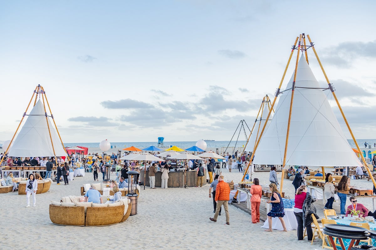 Evénement sur la plage avec des tentes blanches, des parasols colorés et des invités se mélangeant sur le sable près de l'océan.